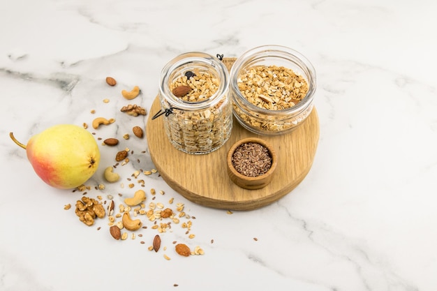 Twee verschillende glazen potten met walnoten granola staan op een ronde snijplank. een marmeren achtergrond met verspreide noten erop en een rijpe peer.