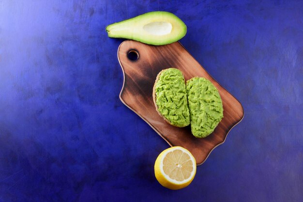 Twee verse groene avocadotoosts op een houten plank Toast een halve avocado en een gele citroen in een schil op een blauwe achtergrond Close-up