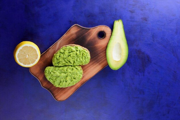 Twee verse groene avocadotoosts op een houten plank Toast een halve avocado en een gele citroen in een schil op een blauwe achtergrond Close-up