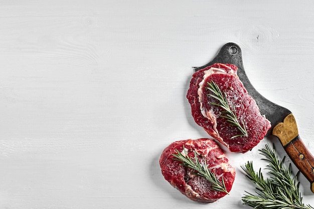 Twee verse rauwe steaks met kruiden knoflook olijfolie peper zout rozemarijn en een keukenbijl op witte achtergrond Bovenaanzicht