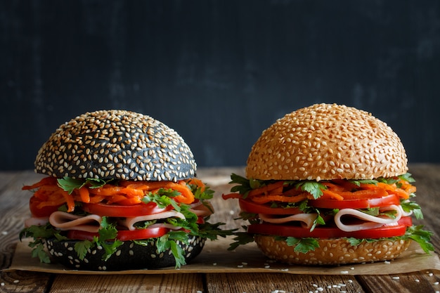 Twee verse smakelijke hamburgers donker en licht met sesamzaadjes, close-up, met verse groenten (tomaat, paprika), Koreaanse wortelen, peterselie en ham. Zwart wazig.