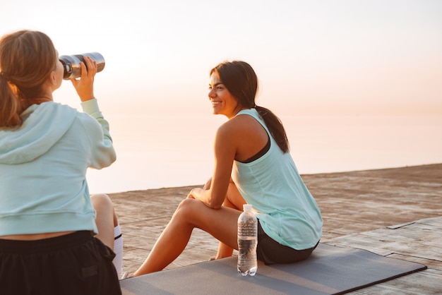 Twee vrienden van sportenvrouwen in openlucht op het strand die en met elkaar zitten spreken