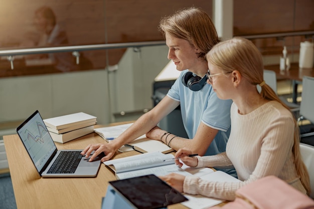 Twee vriendenstudenten die met laptop en boeken in Universiteitsbibliotheek bestuderen