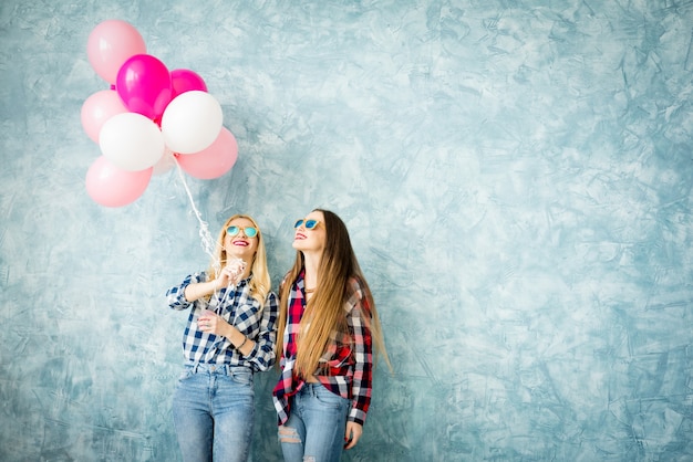 Twee vriendinnen in geruite overhemden die plezier hebben met luchtballonnen op de blauwe muurachtergrond