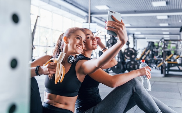 Twee vriendinnen in sportieve kleding zijn in de sportschool fruit aan het luisteren en nemen een selfie.