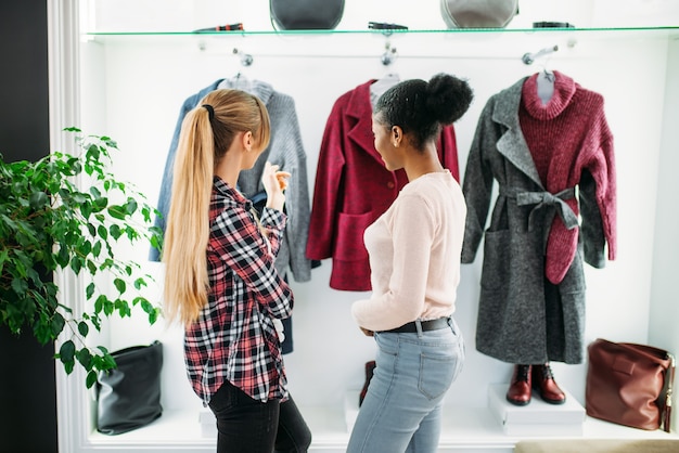 Twee vriendinnen kijken naar de jas in de winkel. Shopaholics in kledingwinkel, levensstijl van consumentisme, mode