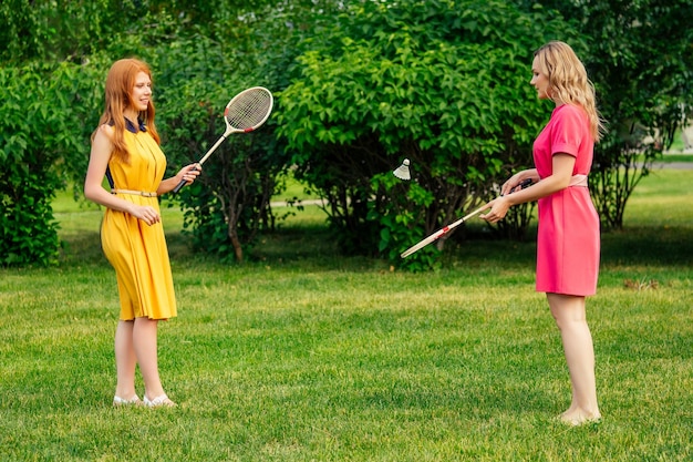 Twee vriendinnen mooie jonge gember roodharige Ierse vrouw in een gele jurk en Europese blonde vrouwelijke persoon in een roze jurk tennis badminton racket spelen in het zomerpark