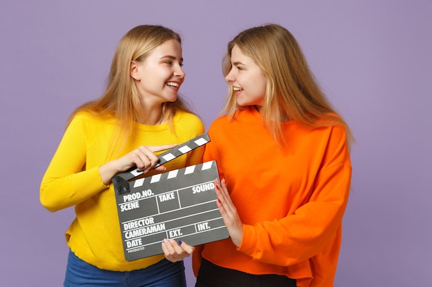 Twee vrolijke jonge blonde tweelingzusters meisjes in kleurrijke kleding met klassieke zwarte film filmklapper geïsoleerd op violet blauwe muur. mensen familie levensstijl concept.