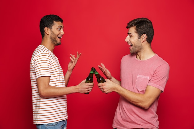 Twee vrolijke jonge mannen staan ?? geïsoleerd over rode muur, frisdrank drinken uit flessen