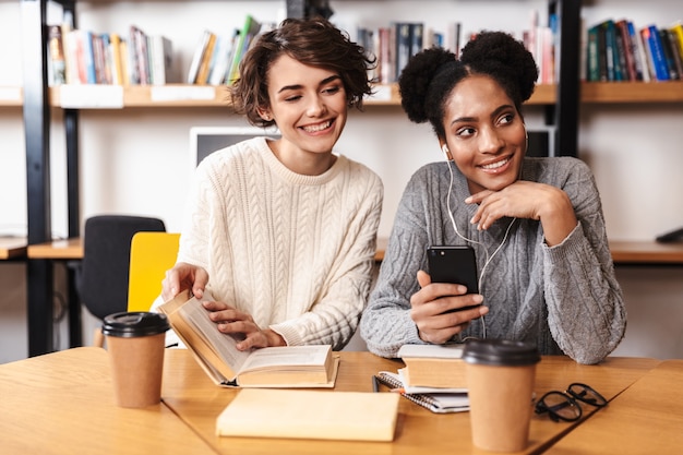 Twee vrolijke jonge meisjes studenten studeren aan de bibliotheek, met mobiele telefoon,