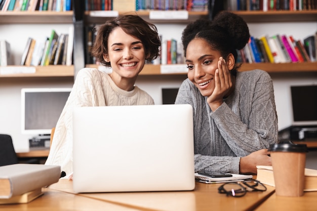 Twee vrolijke jonge meisjes studenten studeren aan de bibliotheek, zittend aan tafel met laptopcomputer