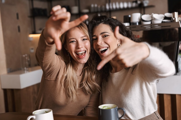 Twee vrolijke jonge meisjesvrienden die aan de cafétafel zitten, samen plezier hebben, koffie drinken