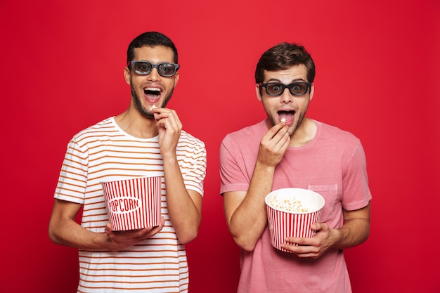 Twee vrolijke jonge mensen die zich geïsoleerd over rode muur bevinden, popcorn eten, 3d bril dragen