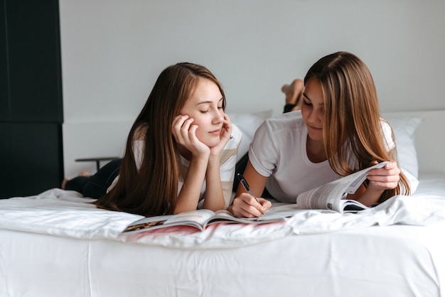 Twee vrolijke jonge tienermeisjes die op bed liggen, tijdschrift lezen