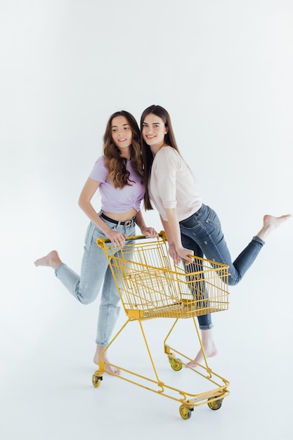 Twee vrolijke jonge vrouwen in kleurrijke trendy outfits die lachen en plezier hebben met trolley