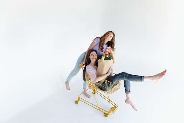 Twee vrolijke jonge vrouwen in kleurrijke trendy outfits die lachen en plezier hebben met trolley