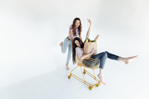 Twee vrolijke jonge vrouwen in kleurrijke trendy outfits die lachen en plezier hebben met trolley