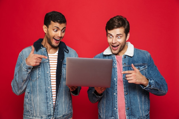 Twee vrolijke mannenvrienden die zich geïsoleerd over rode muur bevinden, laptop computer houden, vieren