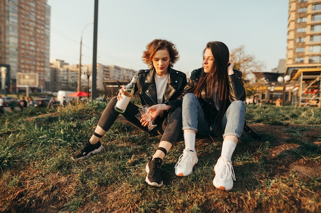 Twee vrolijke meisjes in stijlvolle kleding, zittend op het gras op een achtergrond van stadsgezicht bij zonsondergang