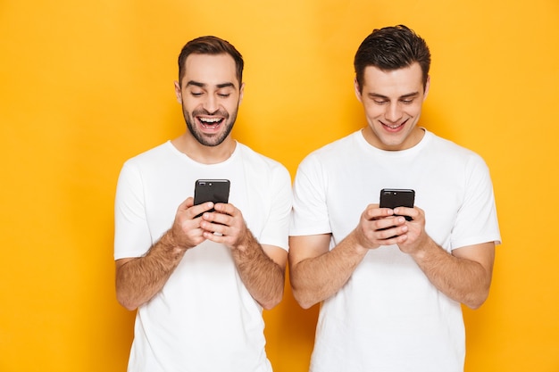 Twee vrolijke opgewonden mannenvrienden die lege t-shirts dragen die geïsoleerd over een gele muur staan, met behulp van mobiele telefoons