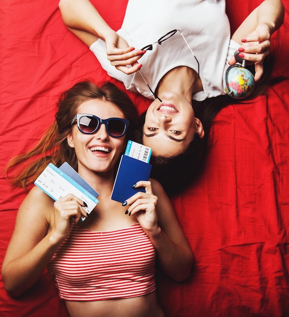 Twee vrolijke vriendenmeisjes die paspoorten, vliegtickets en een wereldbol vasthouden die op een rood bed binnenshuis reisconcept liggen