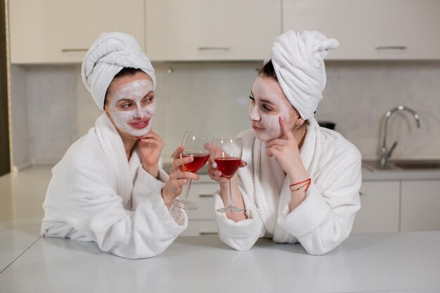 Twee vrolijke vriendinnen in gewaden vieren het weekend drinken rode wijn interieur jeugd en leuk concept foto van hoge kwaliteit