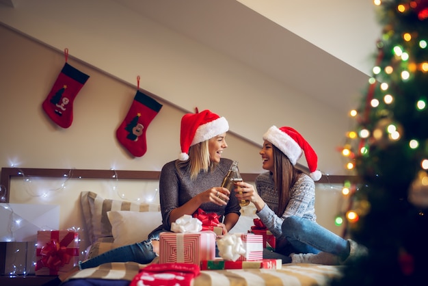 Twee vrolijke vriendinnen met kerstmuts zittend op het bed voor Kerstmis en cheers met bier