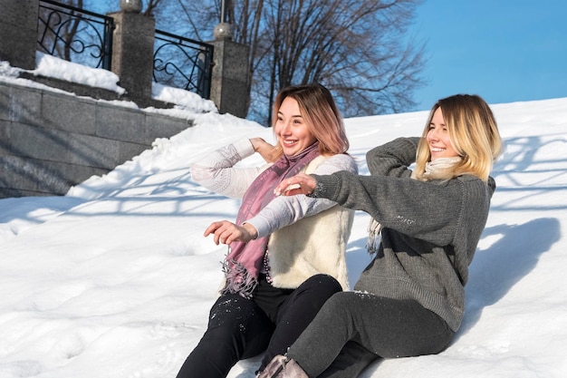 Twee vrolijke vrouwen gooien sneeuwballen zittend in de sneeuw in een stadspark