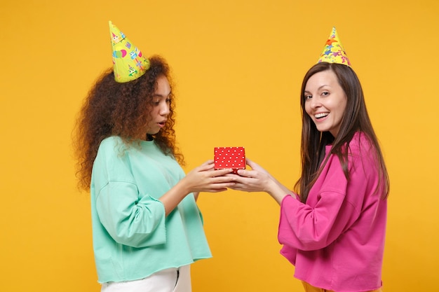 Twee vrolijke vrouwen vrienden europees afrikaans meisje in roze groene kleding verjaardag hoeden poseren geïsoleerd op gele achtergrond. mensen levensstijl concept. bespotten kopie ruimte. houd rode huidige doos met cadeau vast.