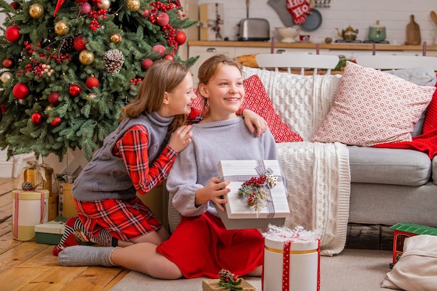 Twee vrolijke zussen zitten thuis bij de kerstboom en kijken naar dozen met cadeaus