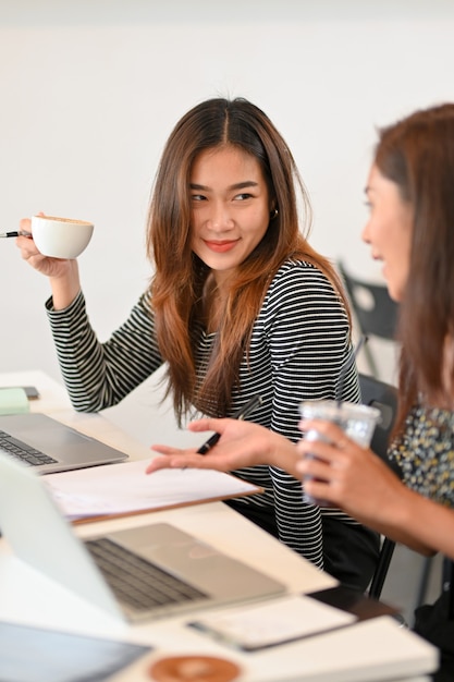 Twee vrouwelijke collega's praten graag tijdens de koffiepauze op kantoor terwijl ze koffie drinken, ontspannende tijd