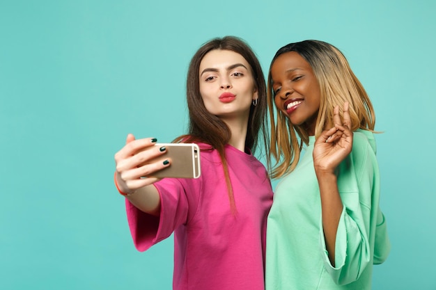 Twee vrouwelijke vrienden Europese en Afrikaanse Amerikaan in roze groene kleding doen selfie schot op mobiele telefoon geïsoleerd op blauwe muur achtergrond, studio portret. Mensen levensstijl concept. Bespotten kopie ruimte.