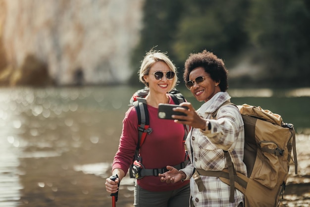 Twee vrouwelijke vrienden van gemengd ras die selfies maken bij een meer tijdens een wandeling langs de berg.