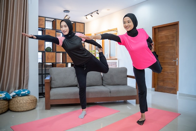 Twee vrouwen die een hoofddoek dragen en glimlachen terwijl ze lunges doen terwijl ze binnenshuis sporten