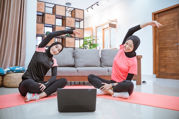 Twee vrouwen die hijab-sportkleding dragen, zitten met gekruiste benen op de grond met hun lichaam naar de zijkant en de handen omhoog, terwijl ze hun armen samen in huis opwarmen