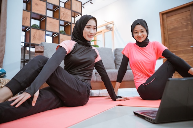 Twee vrouwen die hijab-sportkleding dragen, zitten op de grond terwijl ze hun heupen samen in huis opwarmen