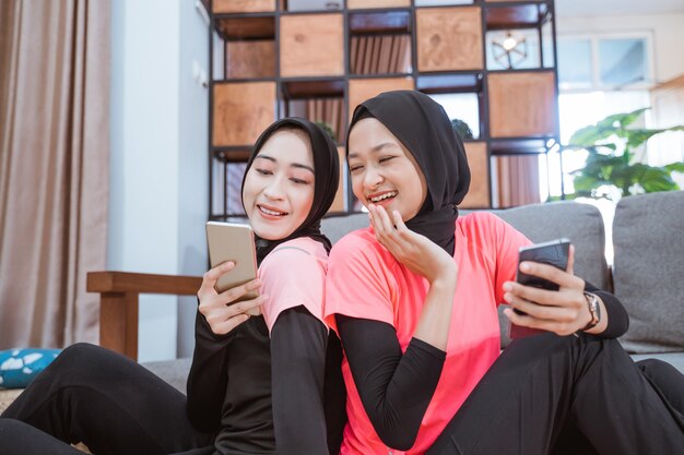 Twee vrouwen die hijabsportkleding dragen, lachen als ze op de grond in huis naar het scherm van een mobieltje kijken