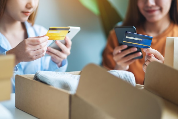 Twee vrouwen die mobiele telefoon en creditcard gebruiken voor online winkelen met boodschappentas en postpakketdoos met kleding op tafel