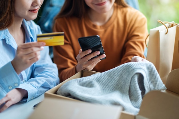 Twee vrouwen die mobiele telefoon en creditcard gebruiken voor online winkelen met boodschappentas en postpakketdoos met kleding op tafel