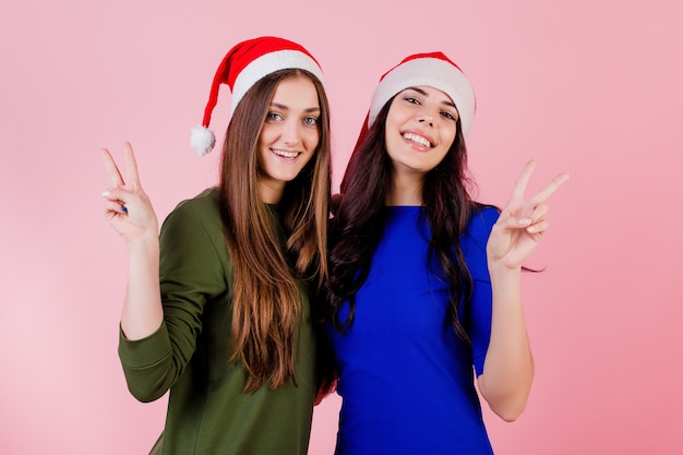 Twee vrouwen die tonend vredesgebaar koesteren die santahoeden dragen die over roze worden geïsoleerd