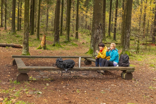 Twee vrouwen lachen, zittend op een bankje in het bos. Drink thee of koffie uit een thermoskan