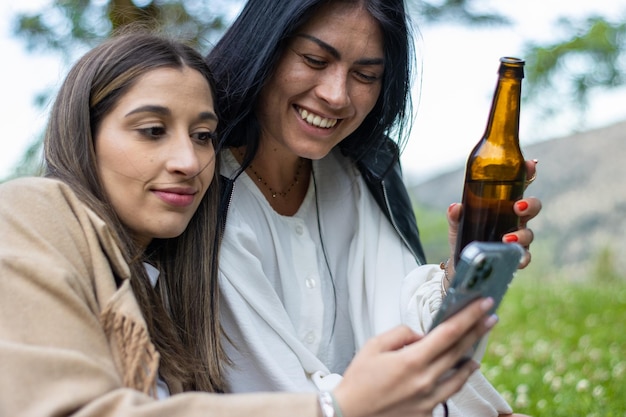 Twee vrouwen met hoeden die op het gras zitten te roosteren met flessen bier terwijl ze hun mobiele telefoons controleren Twee jonge vrouwen met hoeden die op het gras zitten die wijn drinken en hun mobiele telefoons gebruiken