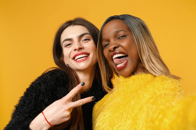 Twee vrouwen vrienden Europese en Afrikaanse Amerikaan in zwarte gele kleding doen selfie shot poseren geïsoleerd op fel oranje muur achtergrond studio portret. Mensen levensstijl concept. Bespotten kopie ruimte