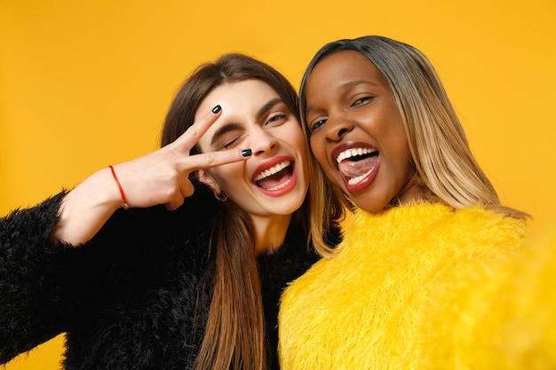 Twee vrouwen vrienden Europese en Afrikaanse Amerikaan in zwarte gele kleding doen selfie shot poseren geïsoleerd op fel oranje muur achtergrond studio portret. Mensen levensstijl concept. Bespotten kopie ruimte