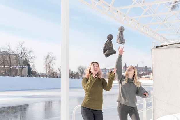 twee vrouwen werpen hun hoed op terwijl ze in de winter aan de oever van een bevroren rivier staan