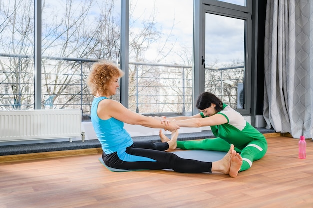 Twee vrouwen zitten met brede benen op de mat en houden elkaars hand vast, in combinatie met stretchoefeningen