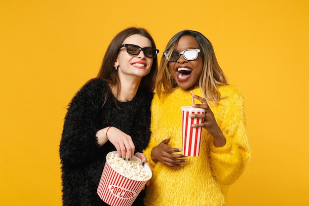 Twee vrouwenvrienden Europese en Afrikaanse Amerikaan in zwarte gele kleren houden emmer popcorn geïsoleerd op een feloranje muurachtergrond, studioportret. Mensen levensstijl concept. Bespotten kopie ruimte.