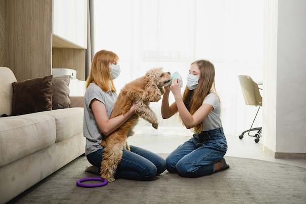 Twee vrouwtjes thuis tijdens de isolatieperiode proberen het mooie huisdier te beschermen tegen virussen, coronavirus, de muilkorf van de hond. Moeder en dochter met een hond.