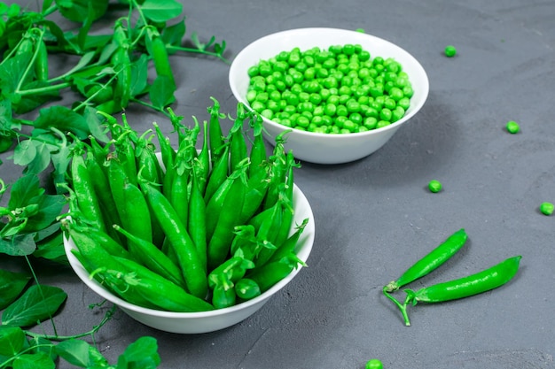 Twee witte kommen verse jonge groene erwten met steken en gepelde bladeren op de achtergrond van scheuten takjes jonge groene erwten op een grijze tafel Close-up Selectieve focus Defocus