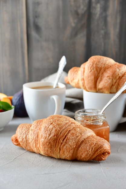 twee witte kopjes koffie met melk, twee croissants en appeljam in een glazen pot